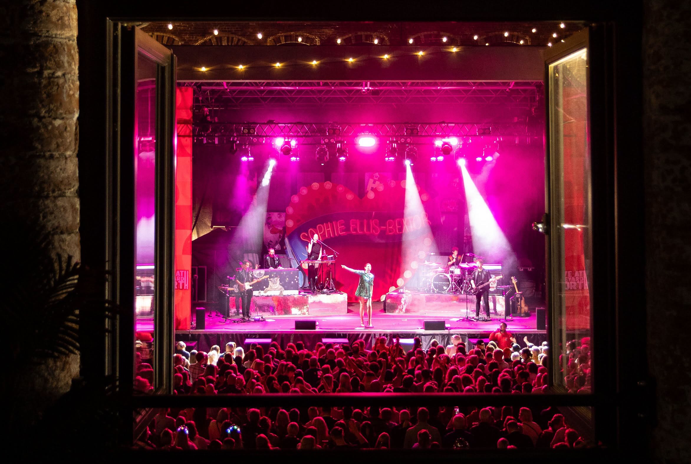 A live band performing on the courtyard stage, framed through an open mill window